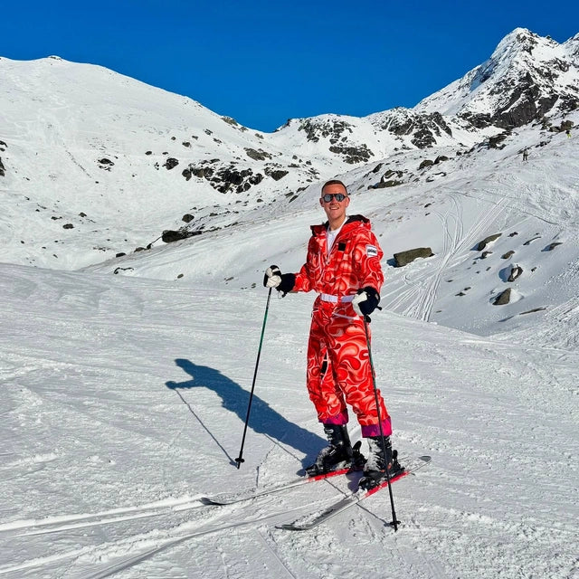 A man wearing a red and orange wavy-patterned ski suit stands on a snowy mountain slope with ski poles, under a clear blue sky.