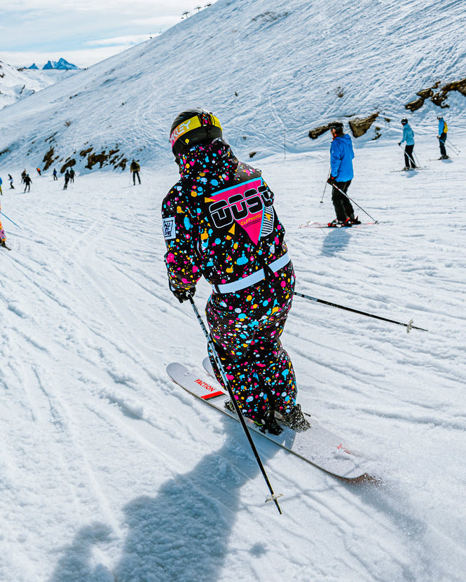 A skier wearing a vibrant, multicolored speckled ski suit with a pink and black Adidas logo on the back, skiing down a snowy mountain slope under a bright sky.