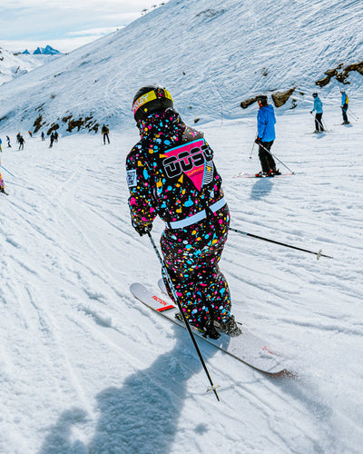 A skier wearing a vibrant, multicolored speckled ski suit with a pink and black Adidas logo on the back, skiing down a snowy mountain slope under a bright sky.