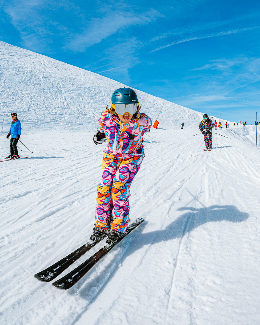 A skier wearing a vibrant, multicolored patterned ski suit and a dark helmet is captured mid-descent on a snowy mountain slope under a bright blue sky. Other skiers are visible in the background.